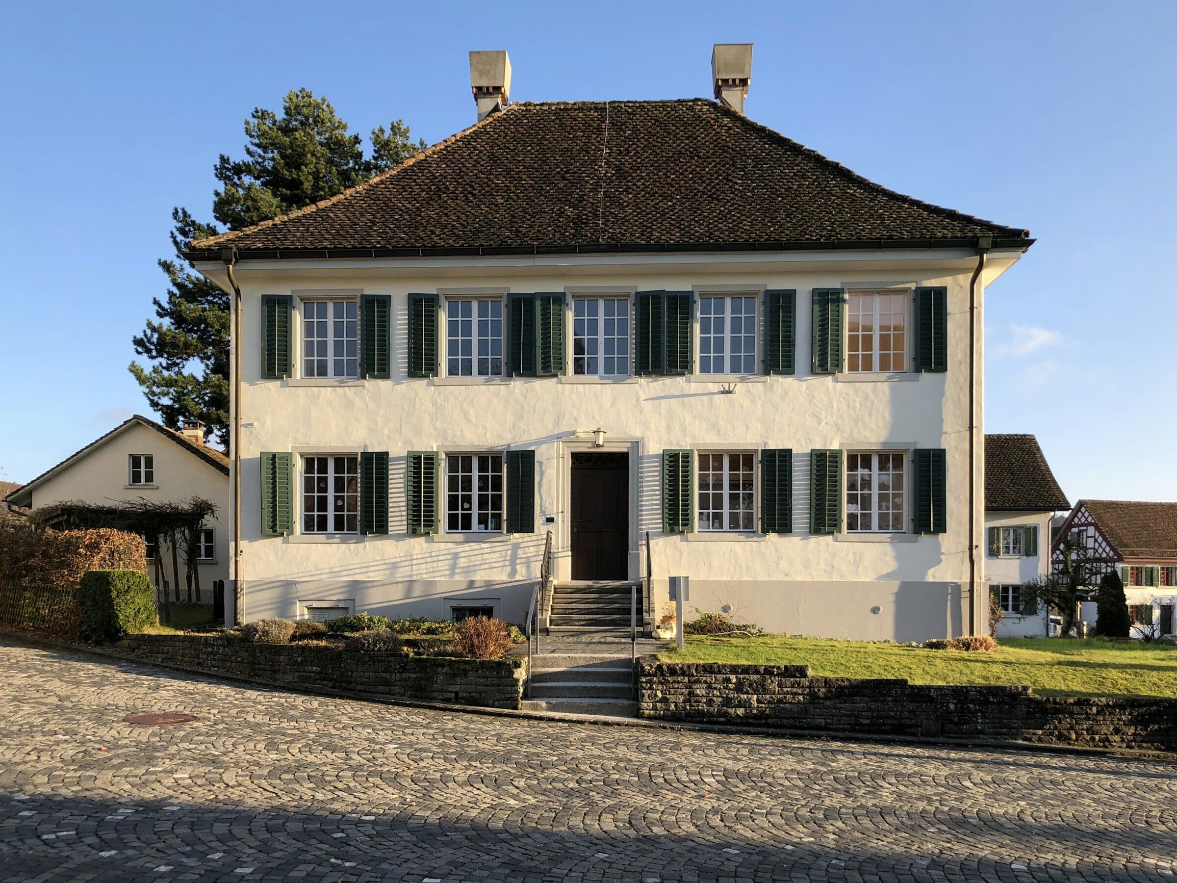 Façade crépie d’une maison de maître, volets verts, au soleil